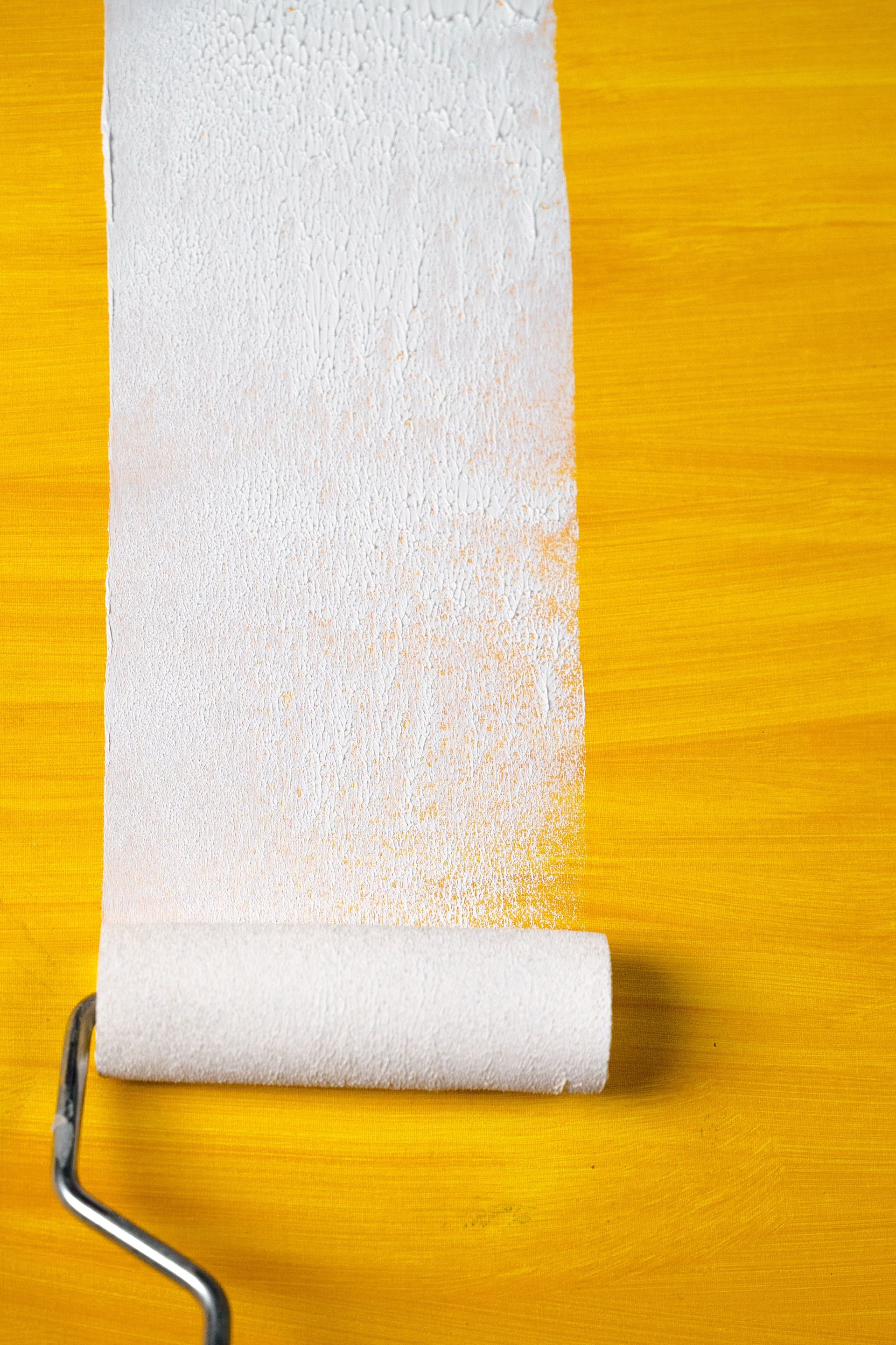 A close-up of a paint roller creating a white stripe on a vibrant yellow surface.